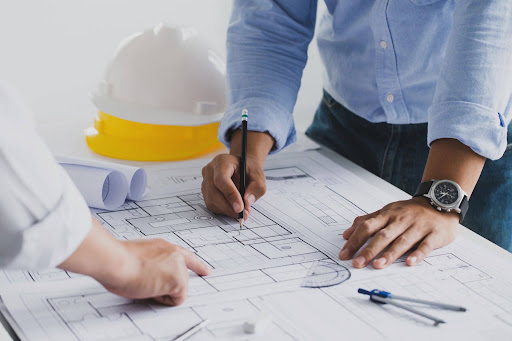Two architects creating a floor plan with two hard hats to the side.